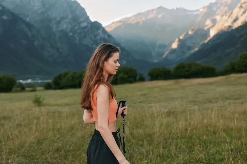 A confident woman stands in an open meadow with trekking poles, gazing toward distant mountains at sunset. She wears a coral top and dark pants, ready for an outdoor adventure.