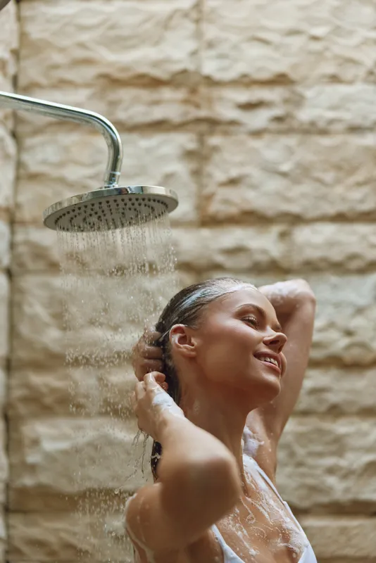 smiling woman washing hair under showerhead with water flowing, enjoying relaxing daily hygiene routine in natural stone bathroom background
