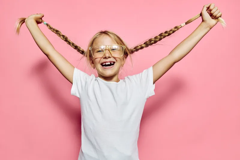 Happy girl in white t-shirt and glasses pulls her braided hair on a pink background. Smile shows missing teeth.