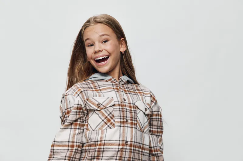 Smiling girl in plaid shirt with long hair posing happily against white background. Child expression with joy and casual style for lifestyle portrait in studio lighting.