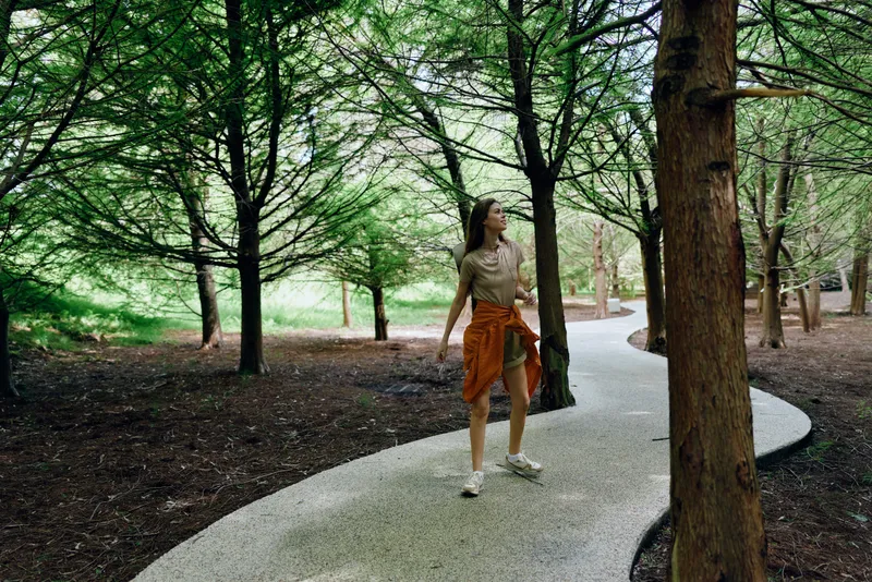 Woman walking on winding path through forest park among tall trees and nature, casual outfit with jacket tied at waist, looking up during a relaxed outdoor stroll.
