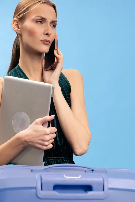 Serious woman holding laptop and talking on phone in front of blue background. Concept of business communication, remote work, travel preparation, and technology use.