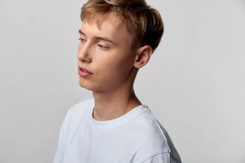 Young man in a white shirt gazes to the side under soft studio light, conveying quiet introspection, calm mood, and contemporary casual style in a clean setting