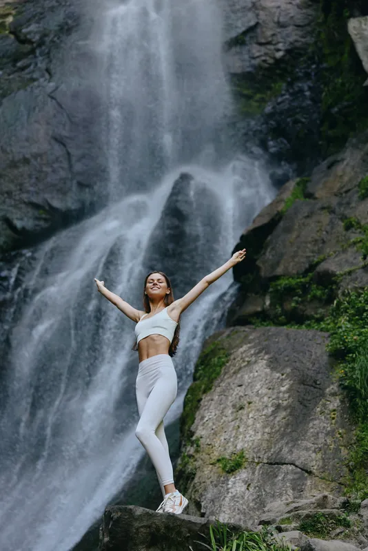 woman practicing yoga in white outfit stands with arms raised near a waterfall, surrounded by rocks and lush greenery in nature