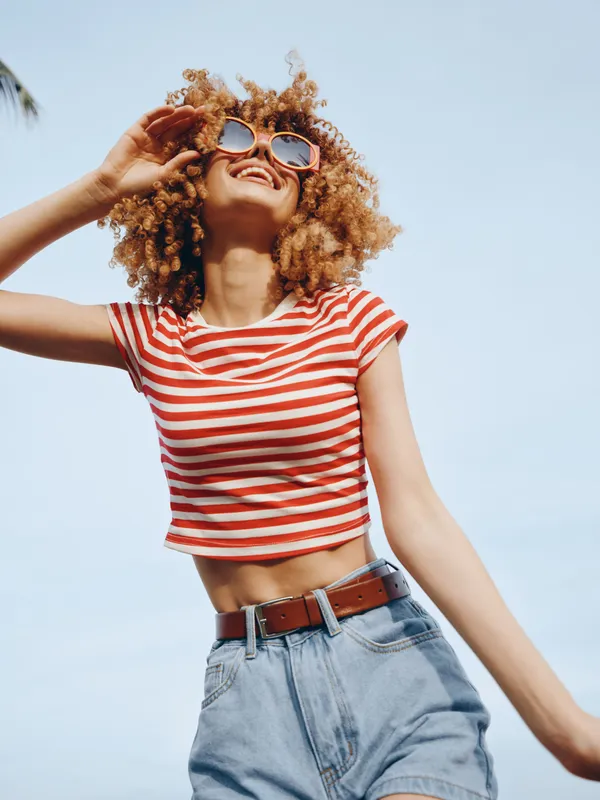 Joyful woman with curly hair wearing a red and white striped crop top and denim shorts, posing against a bright blue sky with carefree energy.