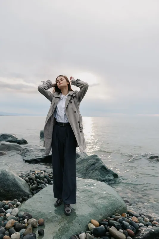 Young woman in a coat stands on rocky beach near calm sea under cloudy sky. She looks relaxed with hands behind head, enjoying peaceful coastal atmosphere and cool weather.