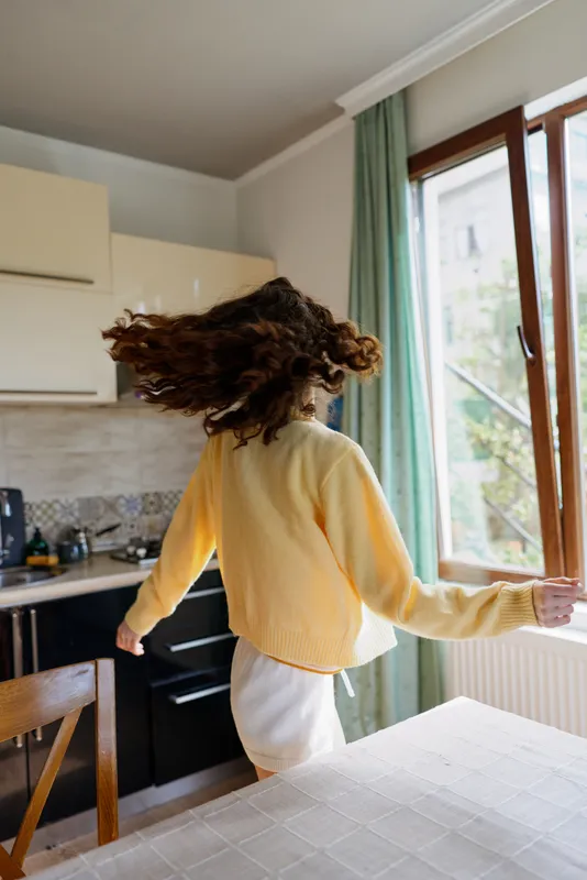 Woman in a cozy home kitchen, spinning with hair in motion, sunlight streaming through a window, casual outfit, candid lifestyle moment, warm colors, relaxed vibe as breakfast light fills the space