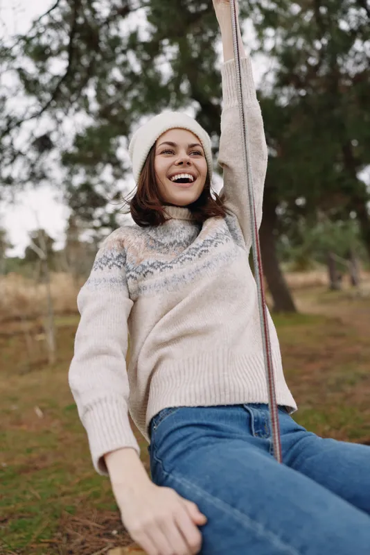 Joyful woman on a swing outdoors in a cozy knit sweater and beanie, laughing as she holds the ropes in a park setting with trees and soft natural light.