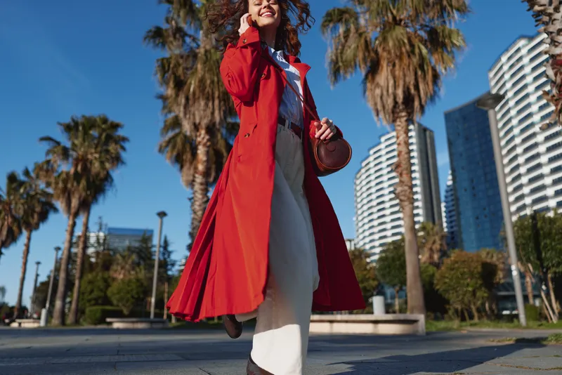 Young woman in a flowing red coat walks confidently in an urban park on a sunny day with palm trees and modern buildings in the background.