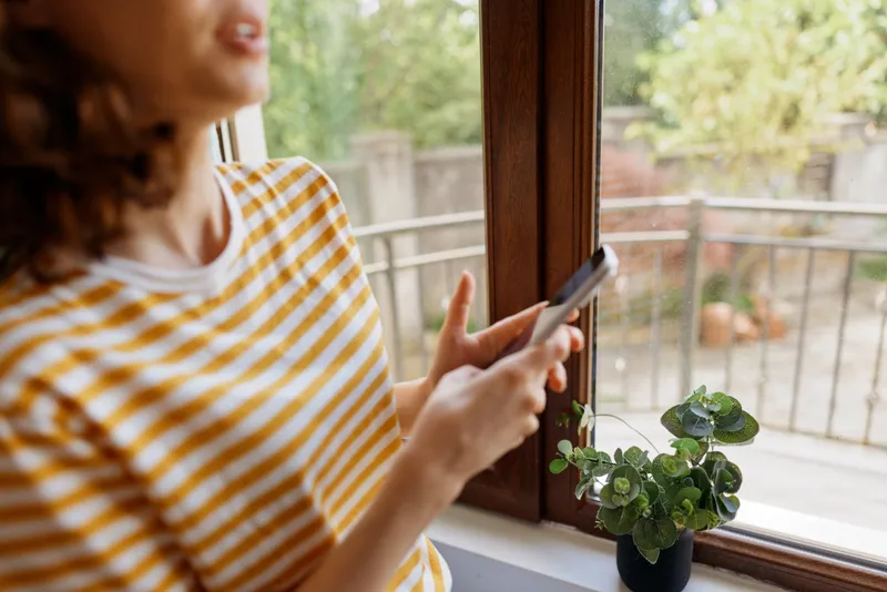 Casual woman with striped shirt using a smartphone by a window, capturing a cozy home lifestyle moment, soft natural light, warm mood, small plant on sill