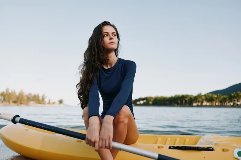A solitary woman sits on a bright yellow kayak at calm waters, wearing a navy long sleeve top, gazing into the distance as sunlight highlights her confident pose.