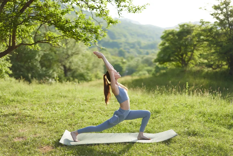 Young woman practicing yoga outdoors in a serene green landscape, wearing blue athletic wear, showcasing a peaceful expression in warm sunlight