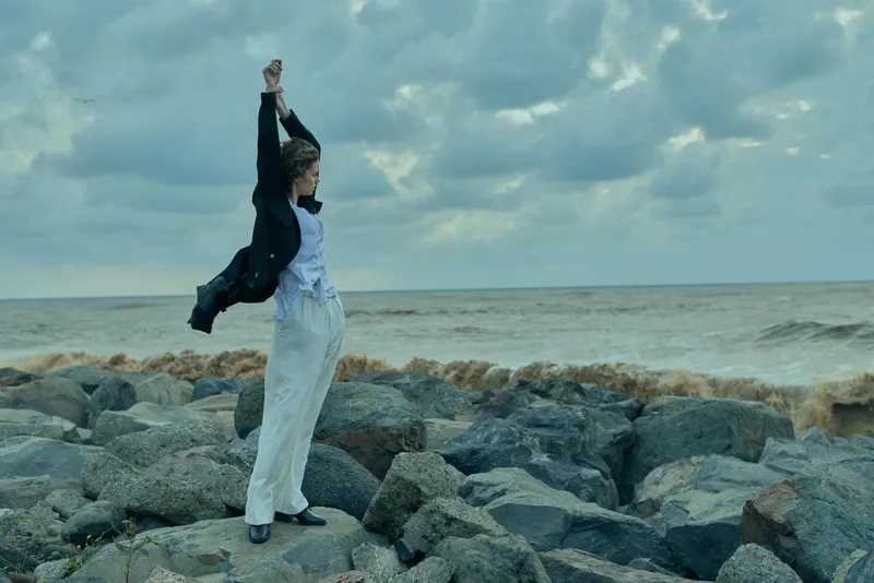 Woman on rocky coast raises arms as wind blows her coat, sea on the horizon, dramatic outdoor scene conveying resilience and freedom by the shoreline.