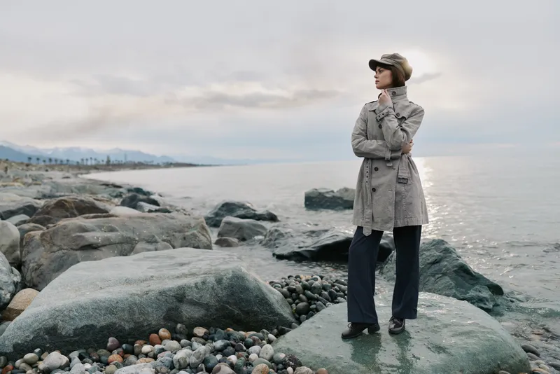 Woman in trench coat and hat stands on rocky shore near calm sea under cloudy sky. Autumn fashion concept with natural landscape and serene mood by water with stones and distant mountains.
