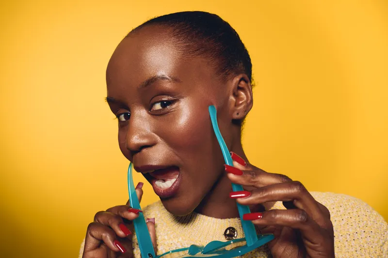 Portrait of cheerful black African woman with dark skin holding blue sunglasses near face on yellow background. Concept for fashion, beauty, and joyful expression in studio lighting.