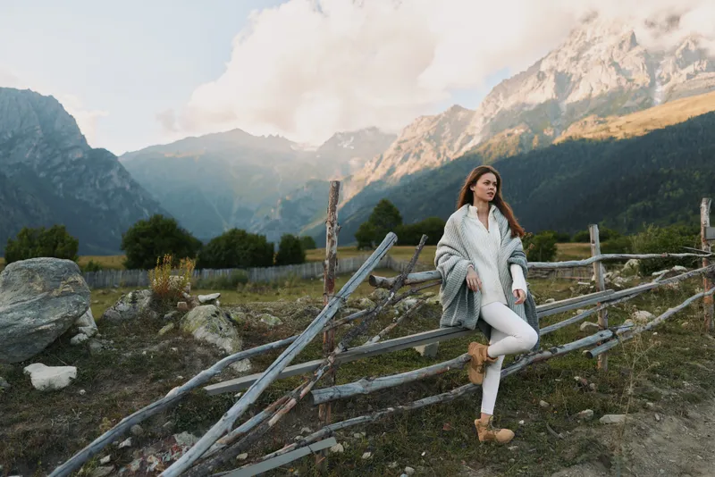 Young woman in a white coat leans against a rustic wooden fence among alpine mountains, calm and reflective in a pristine natural setting with soft light.