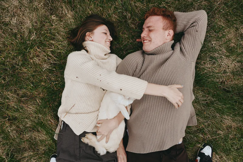 Couple lying on grass enjoying a warm moment with a playful goat kid nearby. Cozy sweaters, natural light, candid lifestyle scene conveying affection, relaxation, and connection in outdoor nature.