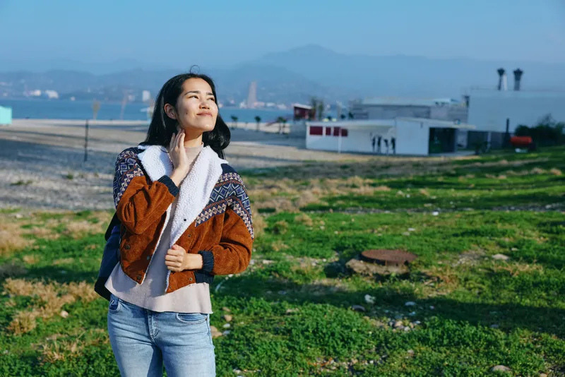 Woman in a cozy scarf and jacket enjoys a sunny outdoor moment by the coastline, with green field, distant buildings and blue ocean under a bright sky, relaxed mood and gentle breeze.