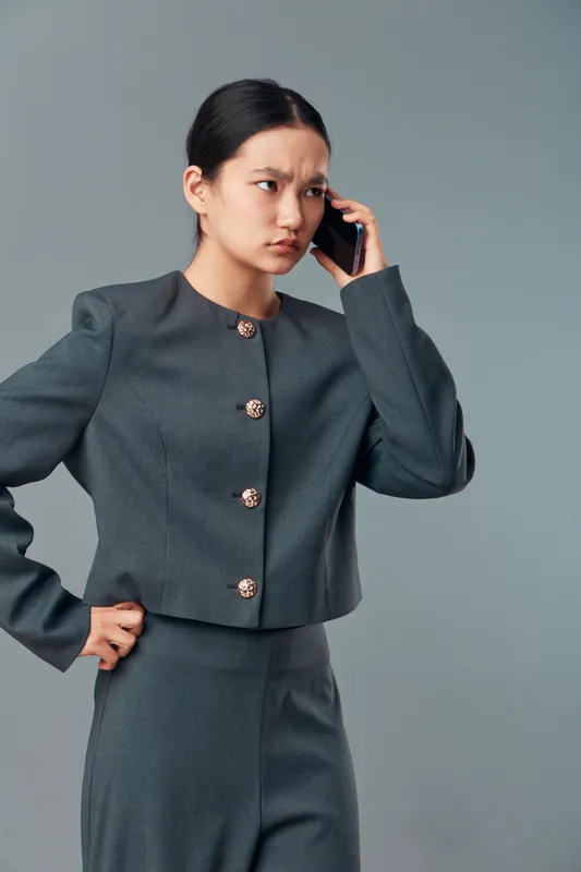 Young woman in business suit looks worried while talking on smartphone. Female professional with serious expression standing on gray backdrop with hand on hip. Communication and emotion.