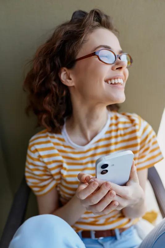 A young woman with glasses laughs while holding a smartphone, wearing a yellow striped tee, seated casually by a bright wall during a relaxed moment in a candid lifestyle setting.
