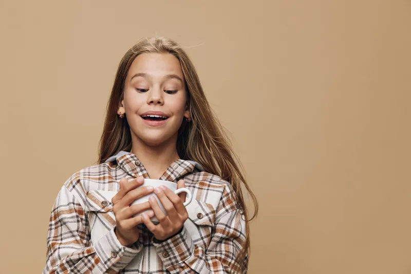 Girl with long hair holding white cup with both hands, smiling and looking down in cozy plaid shirt on beige background, enjoying warm drink in calm atmosphere.