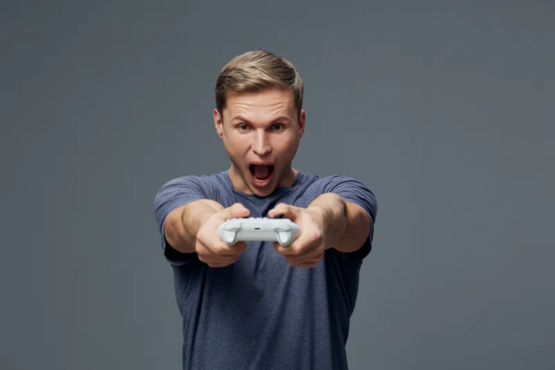 Excited young man playing video game with controller, expressing joy and enthusiasm, casual clothing, gray background, indoor setting, lifestyle concept, entertainment, gaming.