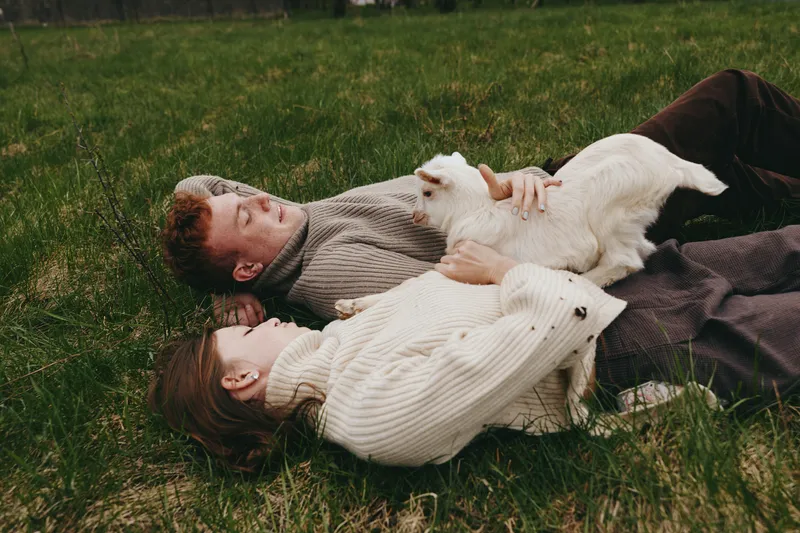 Couple and a playful goat kid lie together on a grassy field, sharing warm affection in soft daylight. A candid lifestyle moment showing relaxation, connection, and natural outdoor comfort.