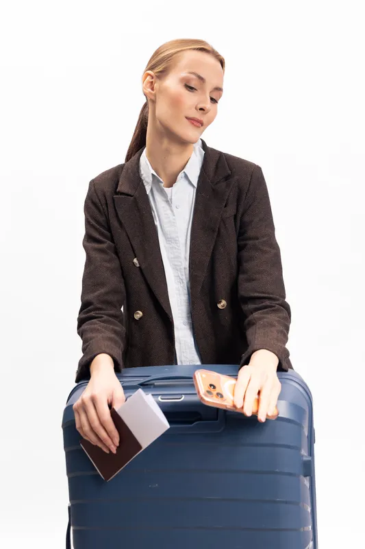 Young woman in blazer holds passport and smartphone while leaning on large suitcase. Travel concept with business casual style and preparation for trip on white background.