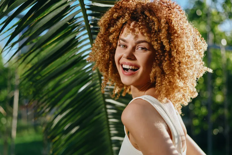 Smiling woman with curly hair enjoys tropical sunlight in white tank top, surrounded by green palm leaves, expressing happiness and natural beauty outdoors.