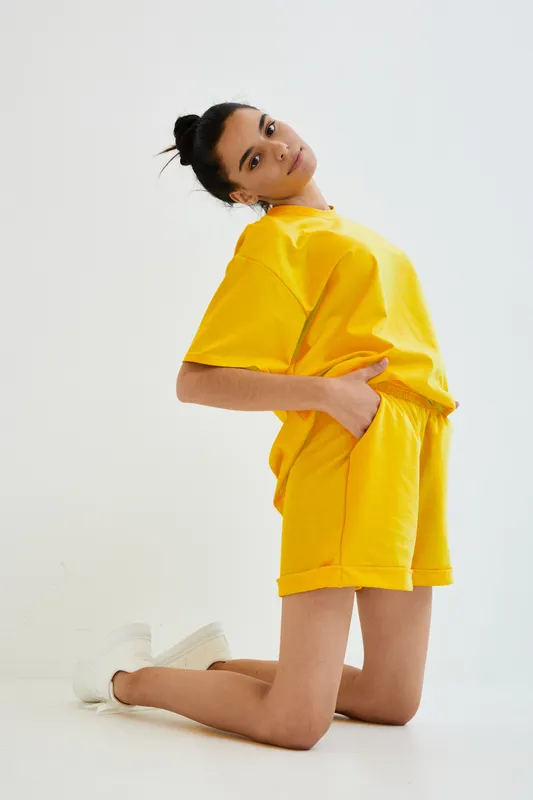young woman wearing yellow casual outfit kneeling on floor with hands in pockets against plain white background, modern fashion style, relaxed confident pose