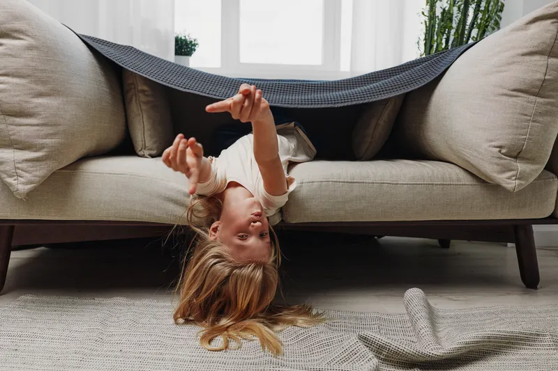 Playful child with long hair hanging upside down from sofa in cozy living room, reaching out with hands. Concept of childhood fun, home leisure, and carefree moments indoors.