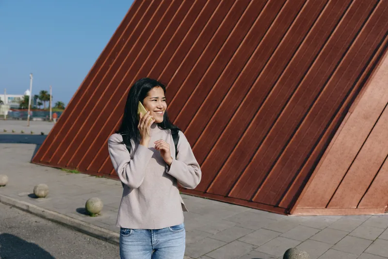 Smiling woman on a phone call outdoors beside bold geometric architecture, casual jeans and sweater, capturing an urban lifestyle moment of communication and everyday chat