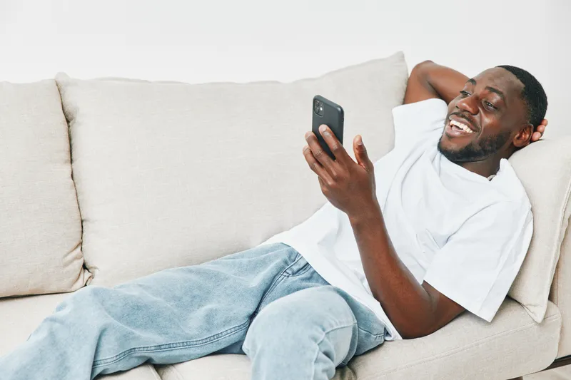 Young African man smiling while using smartphone on a cozy couch, casual attire and relaxed expression Perfect for lifestyle and technology themes