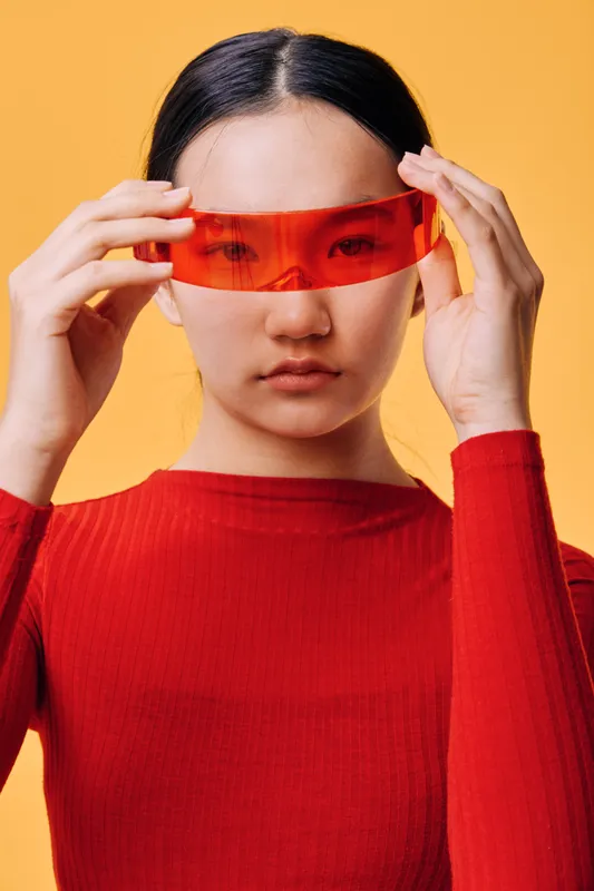 Woman in red sweater adjusting orange translucent futuristic glasses on face with serious expression standing against yellow background for fashion style and technology look.