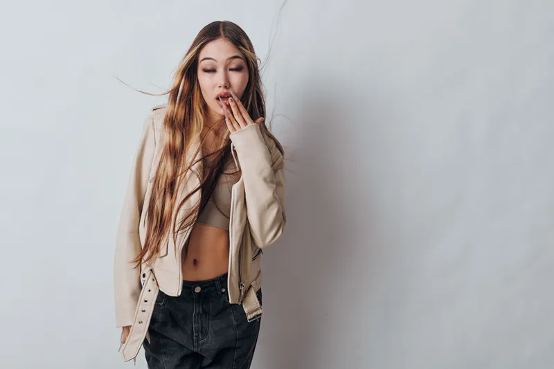 Young woman with long hair wearing beige jacket and crop top covering mouth as if surprised or shocked standing against plain light background in casual style.