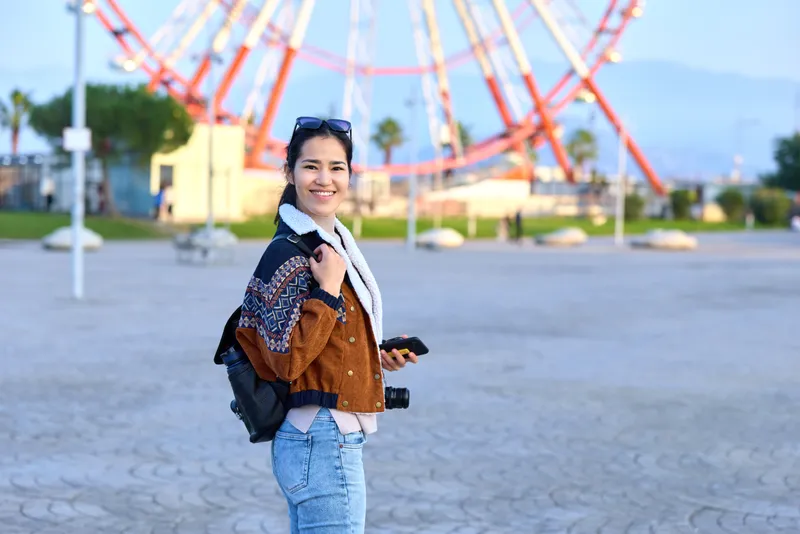 Smiling traveler with backpack and camera at an outdoor park, ferris wheel in background, casual layered clothing, sunny day, adventure and exploration