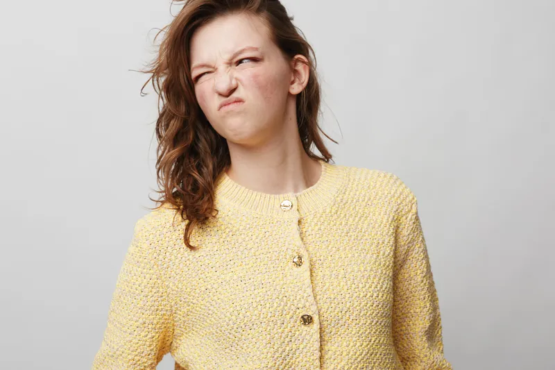 Young woman in yellow knitted sweater makes disgusted face with wrinkled nose and squinted eyes. Isolated on grey background. Concept of dislike, discomfort, and negative emotion.