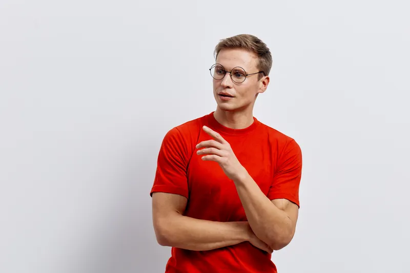 Confident young man with glasses wearing red t-shirt standing against plain background, thoughtful expression, casual style, indoor shot. People lifestyle concept