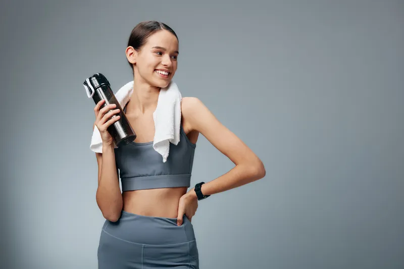 Fit young woman in matching athletic set holds a water bottle and towel, smiling after workout. Minimal studio background with soft lighting highlights toned core and activewear.