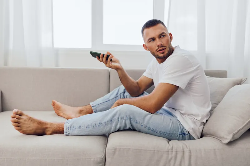 Thoughtful man relaxing on a couch, holding a smartphone, with a modern interior and natural light. Perfect for capturing moments of leisure and reflection.