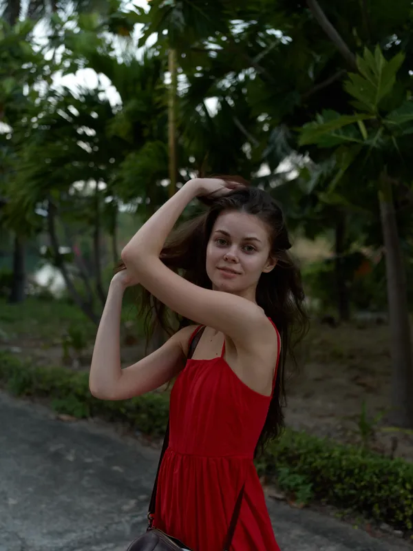 Smiling woman in red dress posing outdoors under green trees during summer evening, natural beauty, casual mood, relaxed atmosphere.