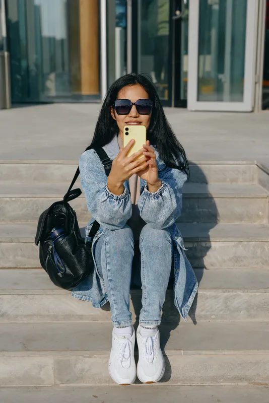 Woman in denim and sunglasses sits on outdoor stairs, holding smartphone with backpack beside, showcasing casual urban fashion and lifestyle accessories, relaxed pose