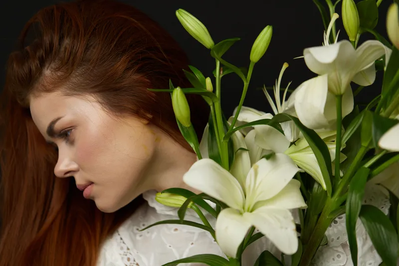 Young woman with long hair holding white lilies, showcasing beauty and elegance in a soft, serene portrait against a dark background