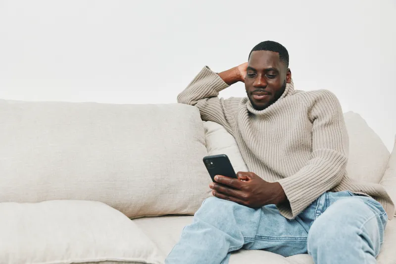 Young Black man relaxing on a couch while using a smartphone, casual clothing, neutral background, modern lifestyle, and comfort style