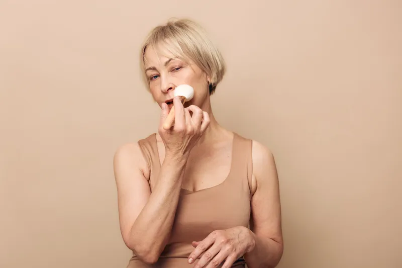 Mature woman with short blonde hair applies powder makeup on face, wearing beige tank top. Minimalist studio portrait with soft neutral background and natural beauty concept.