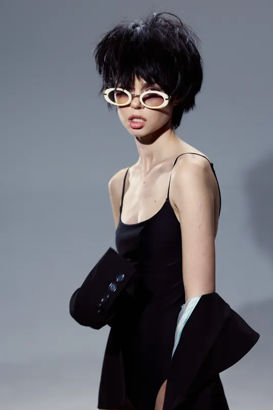 Stylish woman with short hair wearing sunglasses and a black dress against a neutral background, expressing attitude and fashion in studio lighting.