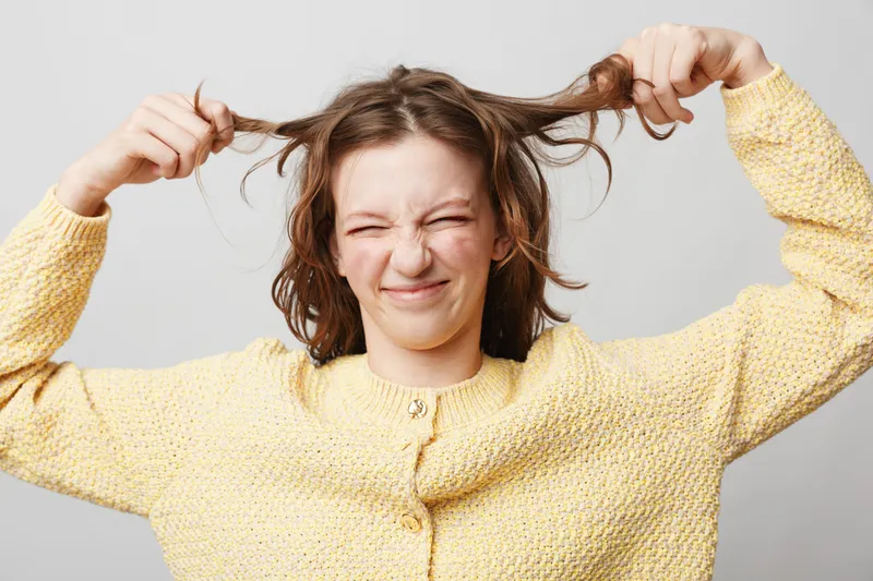 Frustrated young woman in yellow sweater pulls her hair with closed eyes and grimace on face. Isolated on grey background. Concept of stress, anxiety, and emotional tension.