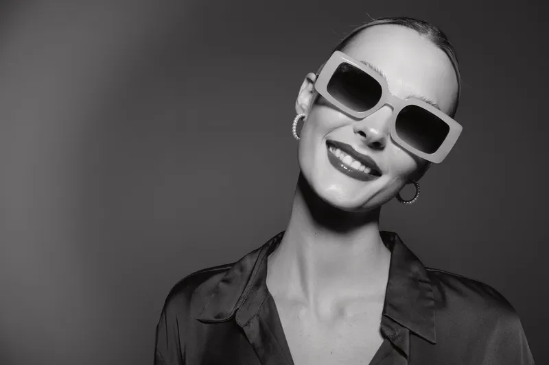 Smiling white woman with pale skin and slicked back hair wearing large sunglasses and hoop earrings. Studio portrait with bright lighting and confident expression.