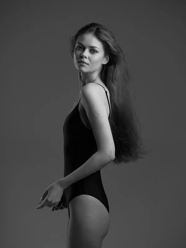 black and white portrait of a young woman in a black bodysuit with long hair, posing confidently against a gray background