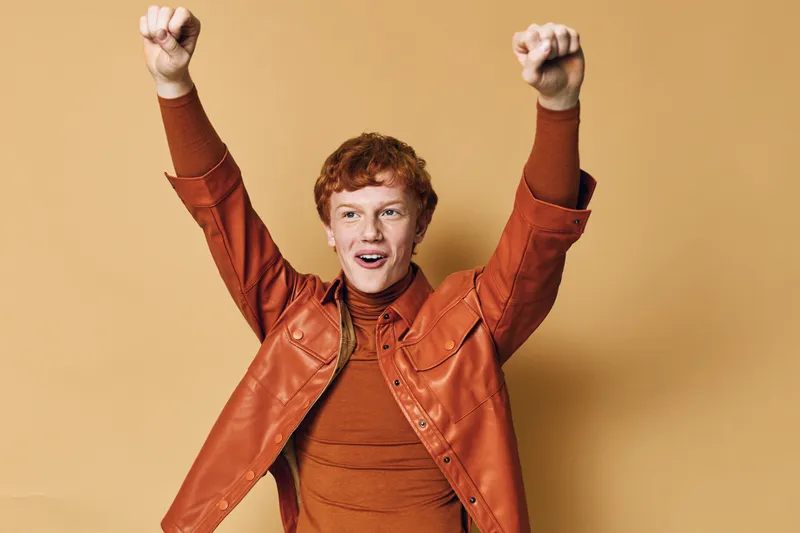 Young man with red hair wearing brown jacket and sweater celebrating with raised fists excited expression on plain beige background.